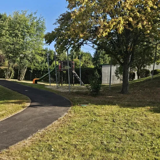 Ein Außenspielplatz mit Rutsche, Klettergerüst und einem gepflasterten Weg. Der Bereich ist von Rasen und Bäumen umgeben. Ein weißes Gebäude ist im Hintergrund zu sehen.