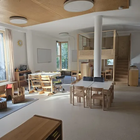 Ein Innenraum eines geräumigen Raumes mit Holzmöbeln, Tischen und Stühlen. Eine weiße Säule steht in der Mitte, und eine Treppe befindet sich rechts. Es gibt Lampen an der Decke, und eine Tafel ist an der Wand befestigt.