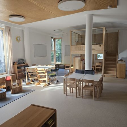 Ein Innenraum eines geräumigen Raumes mit Holzmöbeln, Tischen und Stühlen. Eine weiße Säule steht in der Mitte, und eine Treppe befindet sich rechts. Es gibt Lampen an der Decke, und eine Tafel ist an der Wand befestigt.
