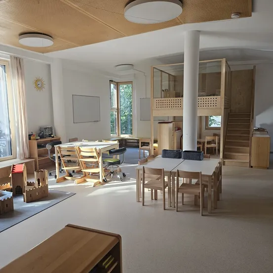 Ein Innenraum eines geräumigen Raumes mit Holzmöbeln, Tischen und Stühlen. Eine weiße Säule steht in der Mitte, und eine Treppe befindet sich rechts. Es gibt Lampen an der Decke, und eine Tafel ist an der Wand befestigt.