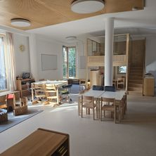 Ein Innenraum eines geräumigen Raumes mit Holzmöbeln, Tischen und Stühlen. Eine weiße Säule steht in der Mitte, und eine Treppe befindet sich rechts. Es gibt Lampen an der Decke, und eine Tafel ist an der Wand befestigt.
