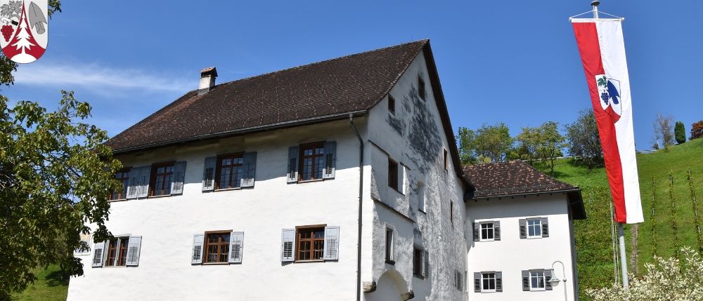 Ein weißes Gebäude mit einem braunen Dach und Fensterläden. Das Gebäude ist von einem Grasfeld mit Bäumen umgeben. Auf der rechten Seite des Bildes befindet sich eine Fahne.