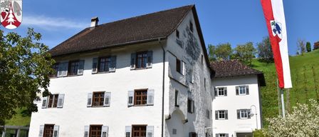 Ein großes weißes Gebäude mit einem braunen Dach, mehreren Fenstern mit Fensterläden und einer Flagge im Vordergrund.