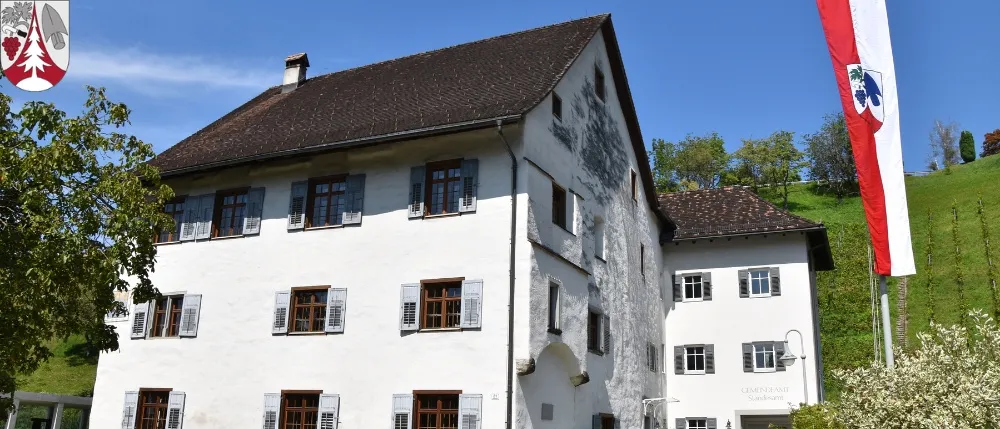 Ein großes weißes Gebäude mit braunen Verkleidungen und Fensterläden, vor dem Hintergrund grüner Hügel und einer Flagge.