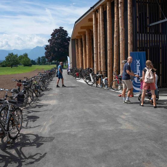 Mehrere Fahrräder sind vor einem modernen Gebäude mit Holzsäulen geparkt. Ein Mann geht, während eine Frau einen Kinderwagen schiebt. Ein blauer Banner steht neben ihnen.