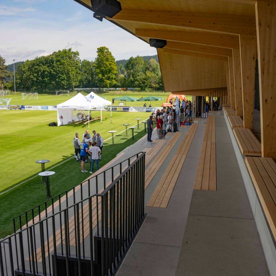 Eine Menschenmenge steht in einer Schlange auf einem Gehweg in der Nähe eines Rasenfelds, möglicherweise in Erwartung einer Veranstaltung. Holzbänke und ein Zaun befinden sich neben dem Gehweg.