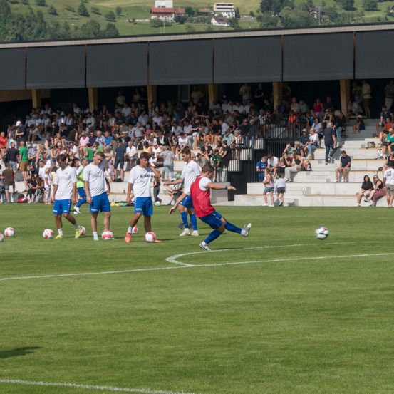 Ein Fußballspiel findet auf einem grünen Feld statt, die Spieler tragen weiße und blaue Uniformen. Zuschauer sitzen auf den Tribünen hinter einer schwarzen Wand.
