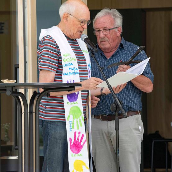 Zwei Männer stehen an einem Podium mit einem Mikrofon, einer hält ein Papier und der andere liest es vor. Sie tragen Freizeitkleidung, und einer hat eine Schärpe mit Handabdrücken.