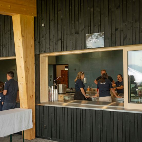 Eine Gruppe von Menschen arbeitet hinter einer Theke in einer Küche. Die Theke hat Edelstahloberflächen. Eine Person trägt ein Shirt mit der Aufschrift 'CREW'. Die Küche hat ein modernes Design mit Holz- und schwarzen Wänden.