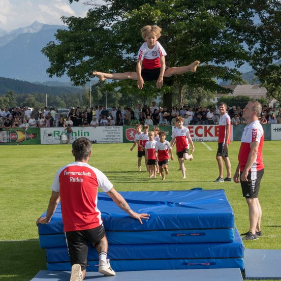 Ein junger Junge in einem rot-weißen Trikot springt über gestapelte Matten auf einem Feld, während mehrere Erwachsene und Kinder dahinter zusehen.