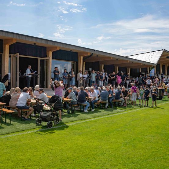 Eine Menschenmenge sitzt auf Bänken auf einem grünen Feld, einige stehend und andere sitzend. Es gibt Tische mit Gegenständen darauf. Ein Mann steht vor einem Podium mit einem Mikrofon.