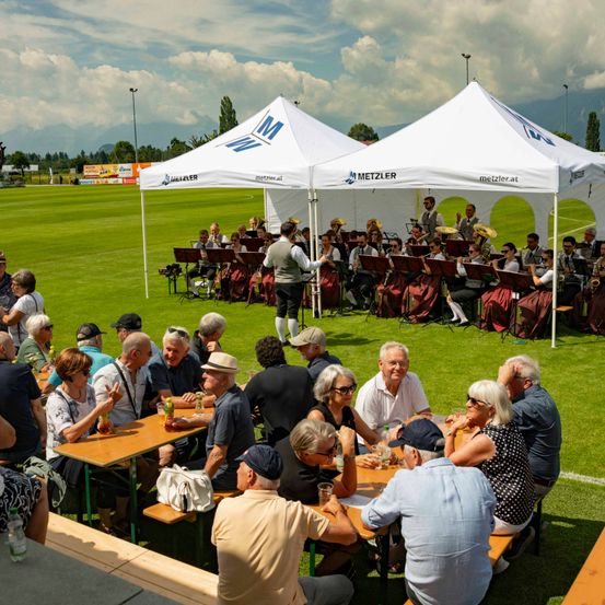 Eine Gruppe von Menschen genießt Getränke und Essen bei einer Veranstaltung im Freien, mit einer Band, die im Hintergrund unter einem Zelt spielt. Der Ort ist auf einem Rasenfeld mit Bergen in der Ferne.