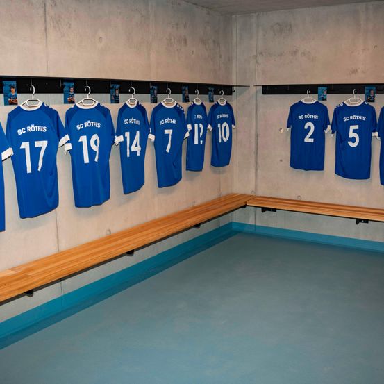 Ein leerer Umkleideraum mit blauen Trikots mit den Nummern 2, 5, 7, 10, 12, 14, 17 und 19, die an einer Wand hängen. Holzbänke säumen den Raum.