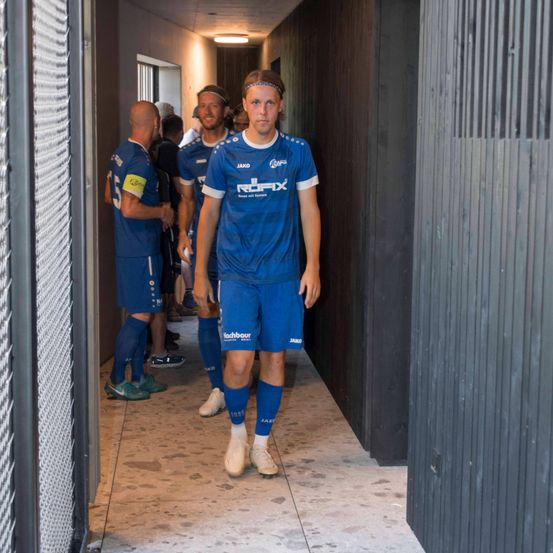 Mehrere Fußballspieler in blauen Trikots gehen durch einen Flur, wahrscheinlich in Richtung Umkleidekabine. Ein Spieler vorne trägt eine Stirnband.