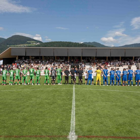 Zwei Fußballmannschaften stehen auf dem Feld, eine in Grün und eine in Blau, mit Zuschauern, die von den Tribünen aus zuschauen.