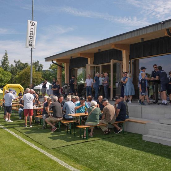 Mehrere Personen sitzen um Tische auf einem grünen Feld, mit einem Gebäude dahinter. Ein Banner steht 'Empire'. Einige Personen stehen herum und andere sind in der Nähe des Eingangs. Bäume und Berge sind im Hintergrund.