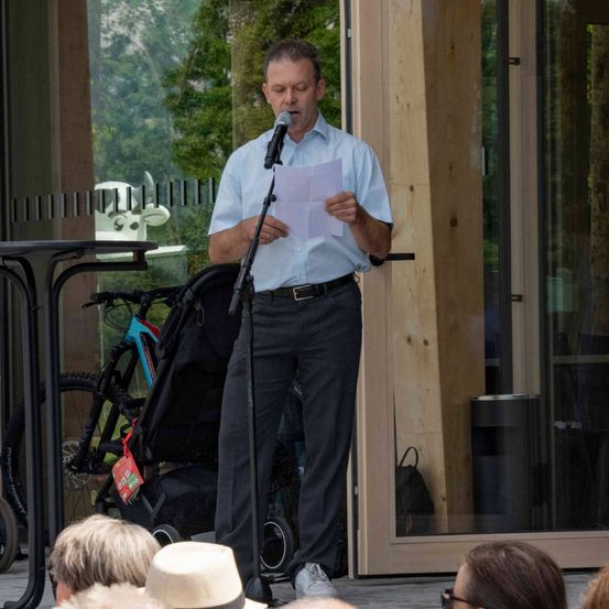 Ein Mann steht auf einer Bühne, hält ein Mikrofon in der Hand und liest von einem Papier vor einer sitzenden Gruppe von Erwachsenen. Hinter ihm stehen ein Kinderwagen und ein Tisch in der Nähe einer Glaswand. Bäume und ein Gebäude sind durch das Glas sichtbar.