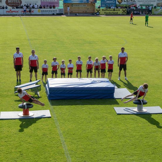Eine Gruppe junger Athleten auf einem Feld, die sich auf eine Leichtathletikveranstaltung vorbereiten. Zwei Personen führen Dehnübungen auf Matten durch, während andere dahinter stehen.
