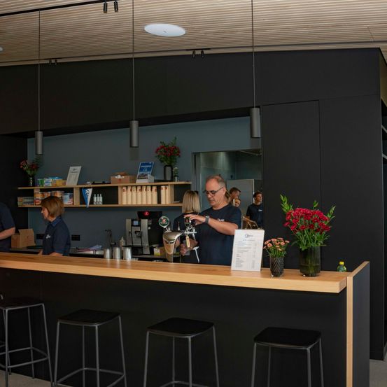 Ein Mann in einem blauen T-Shirt arbeitet hinter einer Theke in einem Café, mit Blumen und einem Menübrett in der Nähe.