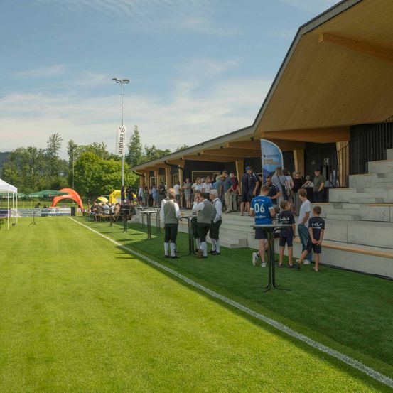Ein Fußballfeld mit einer Menge von Menschen, die stehen und herumlaufen, darunter einige in Trikots. Vor dem Gebäude steht ein Tisch mit einer blauen Fahne.