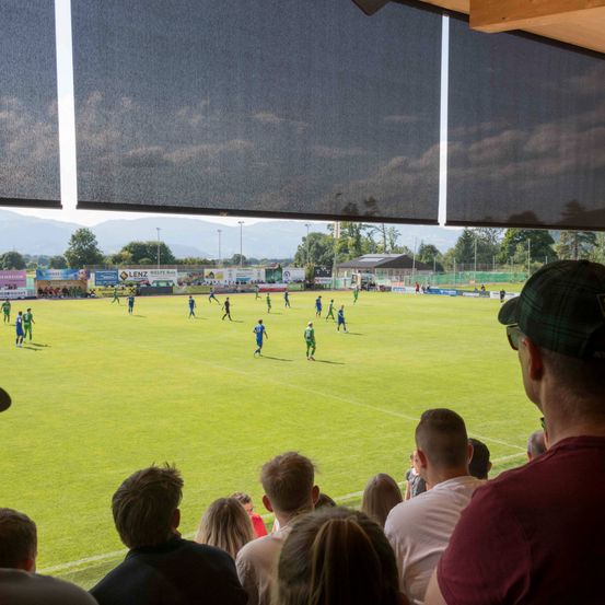 Zuschauer beobachten ein Fußballspiel mit Spielern auf einem Feld, das von großen schwarzen Jalousien beschattet wird. Der Himmel ist teilweise bewölkt, und es gibt Bäume und ein Gebäude in der Ferne.