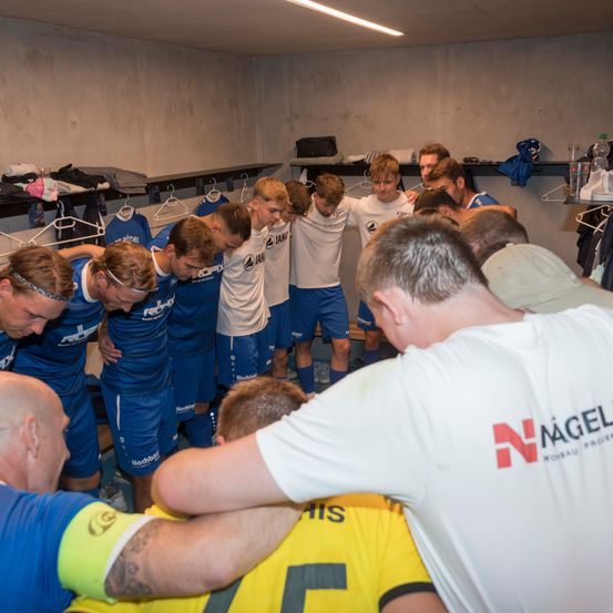 Eine Fußballmannschaft versammelt sich in der Umkleidekabine, mit Spielern in blauen und weißen Trikots, einem in Gelb. Der Trainer, in einem weißen Shirt, führt das Huddle, und der Raum ist mit Sportausrüstung gefüllt.
