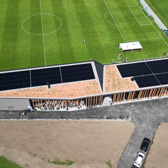 Eine Draufsicht auf ein Fußballfeld mit Solarmodulen auf dem Dach eines Gebäudes. Mehrere Personen sind verstreut und Fahrräder sind geparkt.