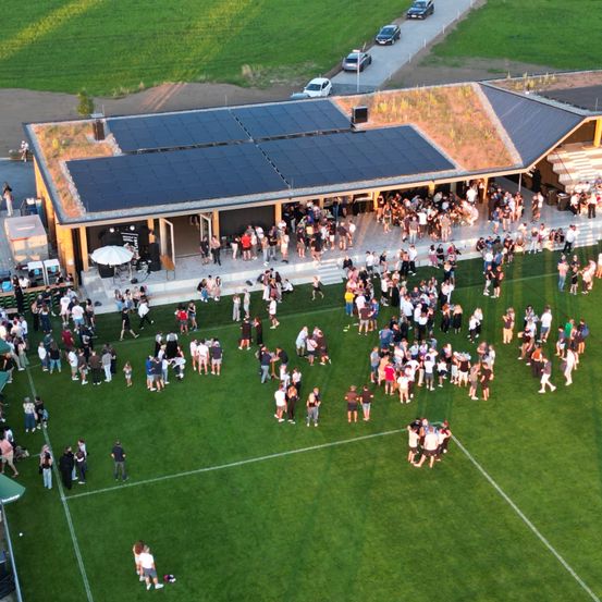 Eine Draufsicht auf eine große Menschenmenge, die sich auf einem Rasenfeld versammelt hat, mit einem Gebäude mit Solarmodulen auf dem Dach im Hintergrund.