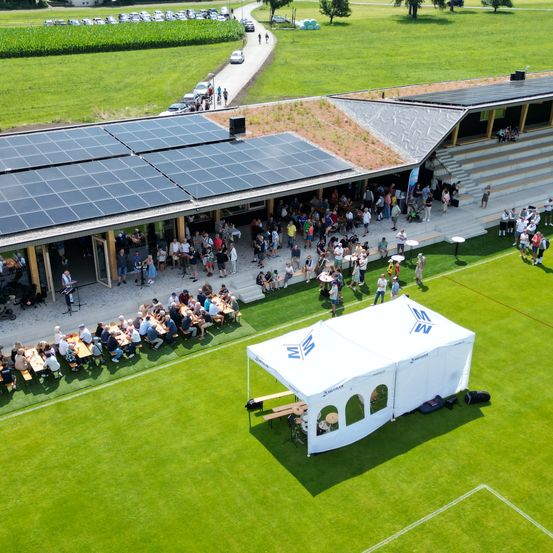 Eine Luftaufnahme einer Sportanlage mit Solarpanelen auf dem Dach. Viele Menschen haben sich für eine Veranstaltung draußen versammelt.