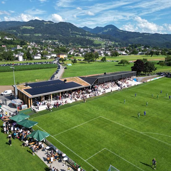 Eine Luftaufnahme eines Fußballfeldes mit einem modernen Gebäude und Solarmodulen, umgeben von einem grünen Feld, Bergen in der Ferne und Menschen, die sich auf den Tribünen versammeln.