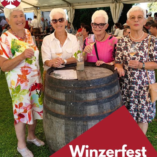 Vier ältere Frauen, alle lächelnd, stehen um einen Holzfass herum mit 'Winzerfest' darauf, Weinläser in der Hand. Sie sind in bunten Kleidern und mit Brillen gekleidet. Der Hintergrund zeigt eine verschwommene Party mit Leuten und Zelten.