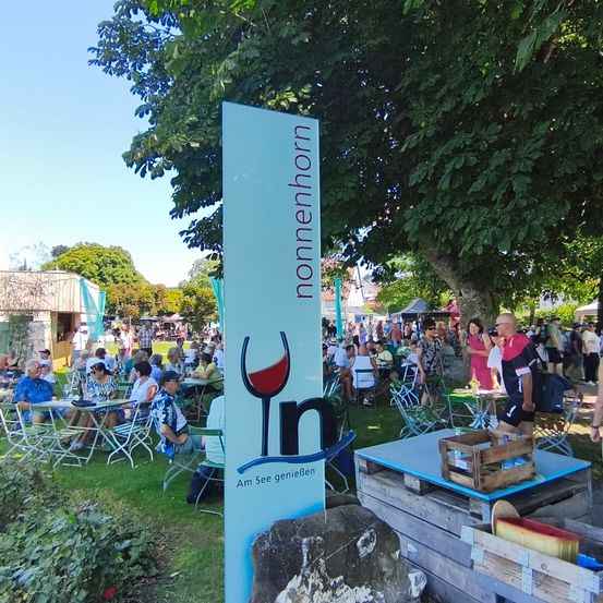 Eine lebhafte Outdoor-Veranstaltung mit vielen Menschen, die an Tischen sitzen und Essen und Getränke unter Bäumen genießen. Ein Schild mit Weinglas und 'nonnenhorn' Logo steht in der Mitte.