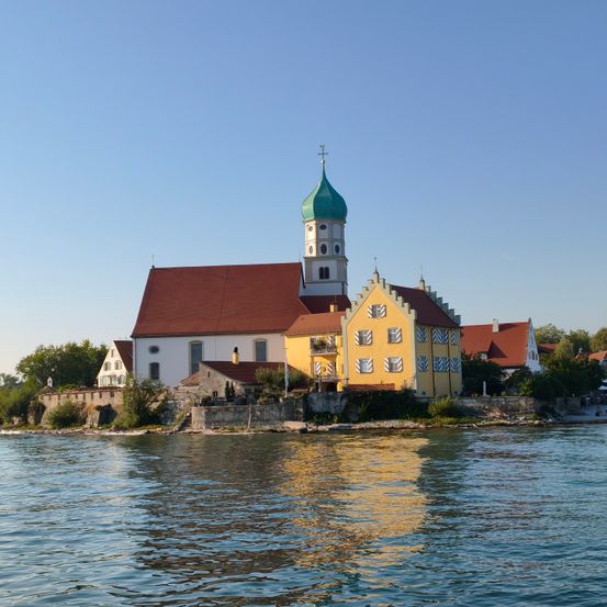 Eine Kirche mit grüner Kuppel und rotem Dach steht auf einer kleinen Insel am Wasser unter einem klaren Himmel.