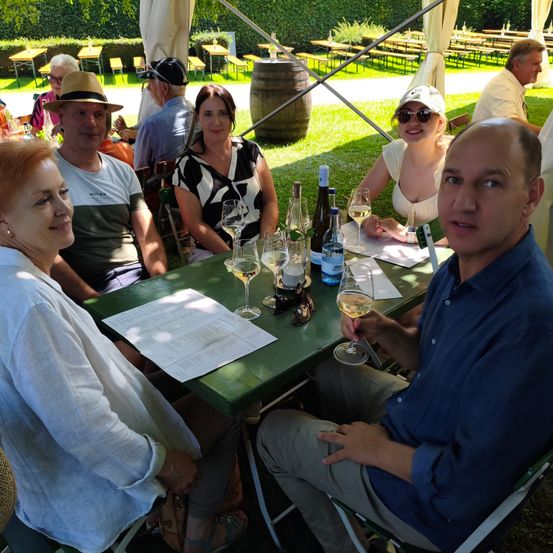 Eine Gruppe von Menschen sitzt um einen grünen Tisch im Freien. Sie trinken Wein und lächeln. Hinter ihnen stehen ein Fass und Tische mit Bänken.