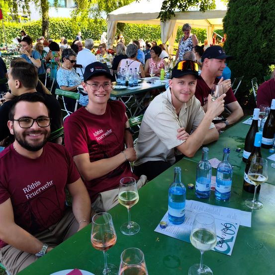 Eine Gruppe von Menschen mit Hüten sitzt an einem grünen Tisch und hält Weingläser in der Hand. Einige lächeln, und der Tisch ist mit Flaschen und einer Speisekarte gedeckt. Bäume und Zelte sind im Hintergrund.