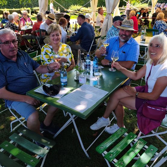 Eine Gruppe von Menschen sitzt um einen grünen Tisch in einem Außenbereich, lächelt und hält Weingläser. Eine Person trägt einen Hut.