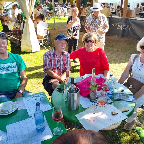 Eine Gruppe von Menschen sitzt um einen grünen Tisch und genießt Drinks. Ein Mann hält ein Weinglas, und der Tisch hat einen Weinkühler und verschiedene Flaschen. Hinter ihnen stehen und sitzen Leute, einige in weißen Zelten.