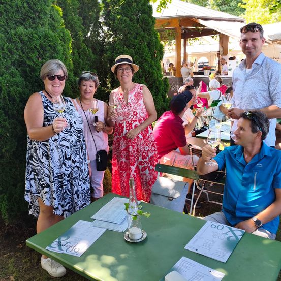 Eine Gruppe von Menschen steht und lächelt bei einer Outdoor-Veranstaltung mit Wein glasses in der Hand. Sie sind in einem Garten mit grünen Pflanzen und einem Tisch mit einem Menü. Hinter ihnen sitzen und stehen weitere Menschen.