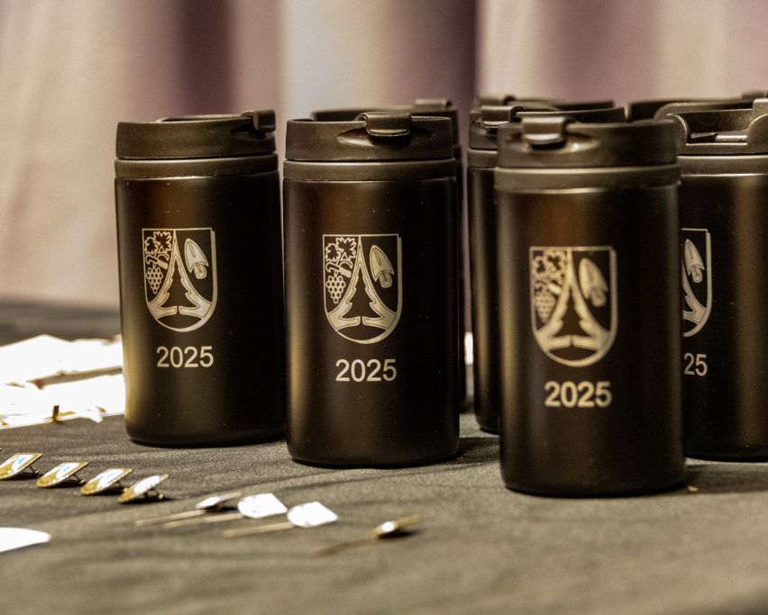 Eine Reihe schwarzer Edelstahl-Trinkbecher mit dem Jahr 2025 und einem Logo sind auf einem Tisch mit Stiften und Abzeichen ausgestellt.