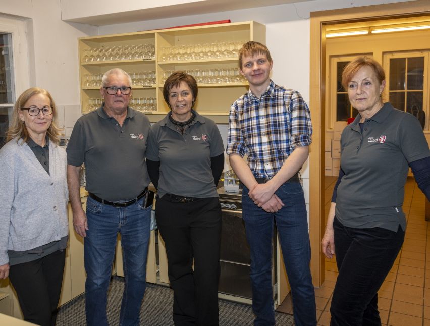 Fünf Personen in grauen Uniformen stehen in einer Küche zusammen. Ein Regal voller Gläser steht hinter ihnen. Der Raum hat geflieste Böden und eine Holztür.