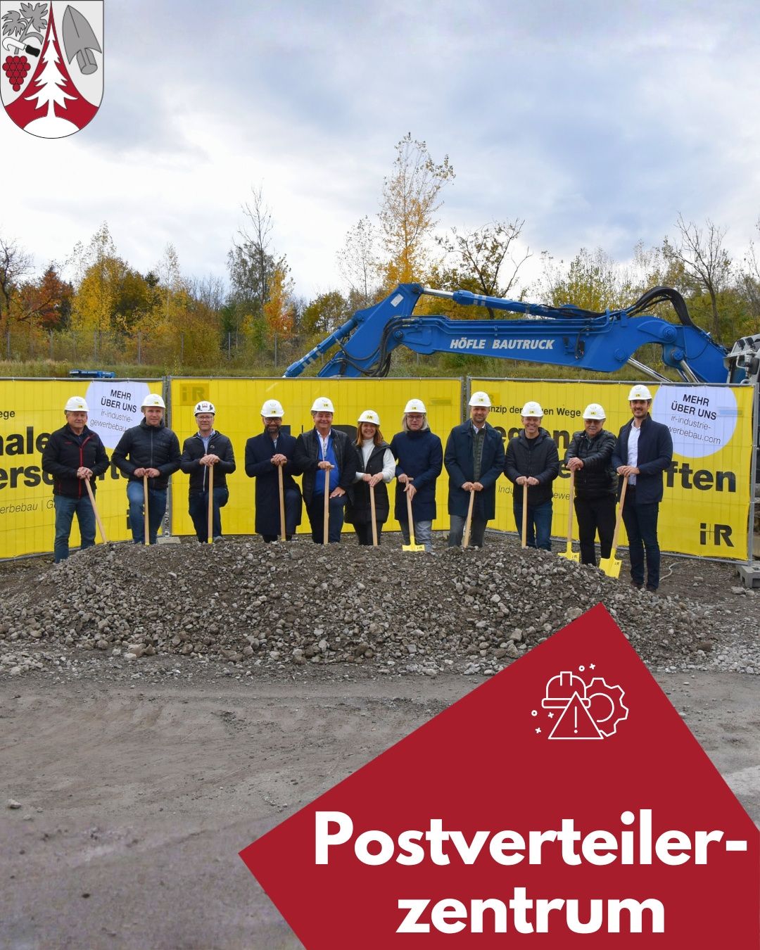 Eine Gruppe von Menschen in Helmen steht vor einer Baustelle mit einem gelben Banner und einem blauen Bagger.