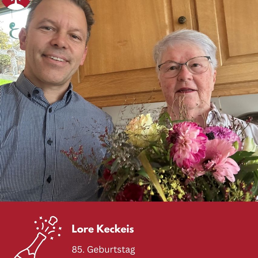 Ein Mann und eine ältere Frau stehen lächelnd in einer Küche. Die Frau hält einen Blumenstrauß. Das Bild hat einen roten Rand und den Text 'Lore Keckeis 85. Geburtstag'.