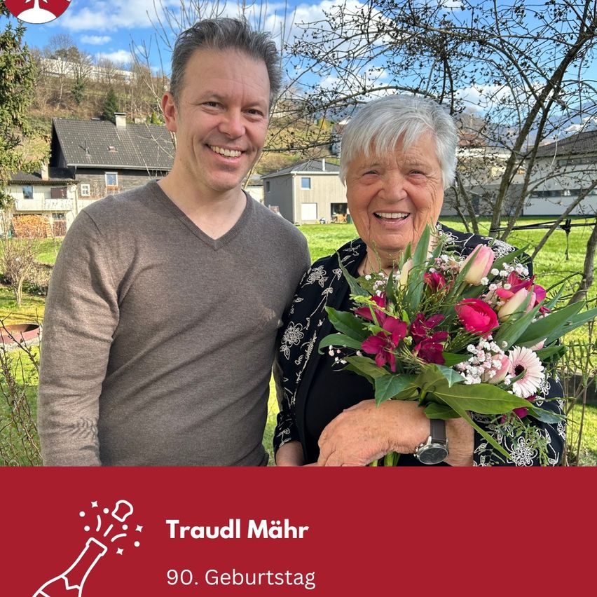 A smiling elderly woman with white hair holds a bouquet of flowers, standing beside a smiling young man in a garden. Behind them are houses and a tree. The banner below says Traudl Mäh and 90. Geburtstag.