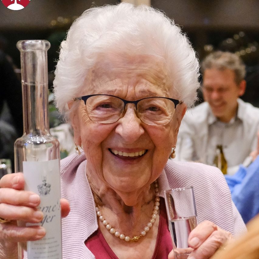 An elderly woman wearing glasses holds a bottle of wine and a glass of liquid, smiling and looking forward. A man in a white shirt looks at something behind her.