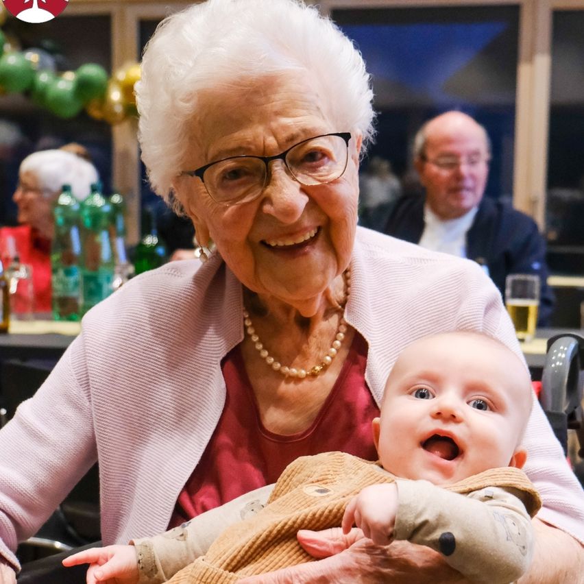 An elderly woman with glasses holds a smiling baby in her arms at a family gathering. Behind them, another man and woman are sitting at a table.