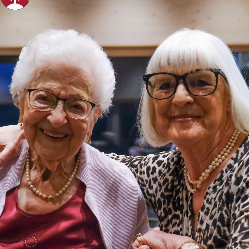 Two older women with white hair, both wearing pearl necklaces and glasses, are smiling and posing for a picture.