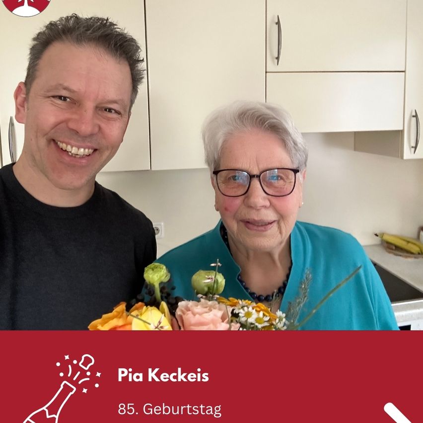 Ein Mann und eine ältere Frau stehen in einer Küche. Der Mann lächelt und hält einen Blumenstrauß. Die Frau trägt eine Brille und eine Halskette. Sie lächeln beide für ein Foto. Hinter ihnen befindet sich eine Arbeitsplatte mit einer Banane darauf.