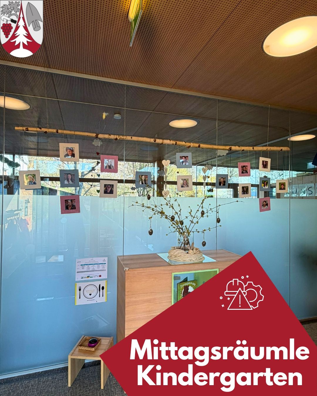 Ein modernes Bürointerieur mit einer Glaswand zeigt verschiedene Fotos und einen dekorativen Zweig. Ein Holztisch hält eine Pflanze und ein rotes Dreieck mit einem Warnzeichen und dem Wort Mittagsraumle.