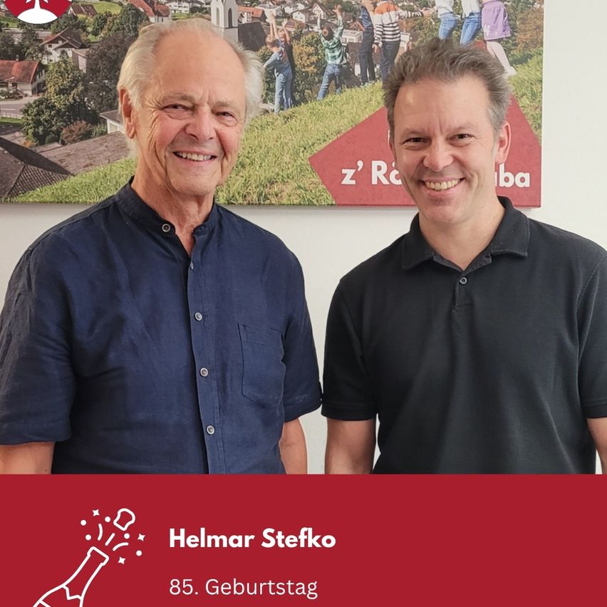 Zwei lächelnde Männer vor einem Poster, einer trägt ein blaues Hemd, der andere ein schwarzes Poloshirt, mit einem darunterstehenden Feierbanner, das 'Helmar Stefko 85. Geburtstag' steht.