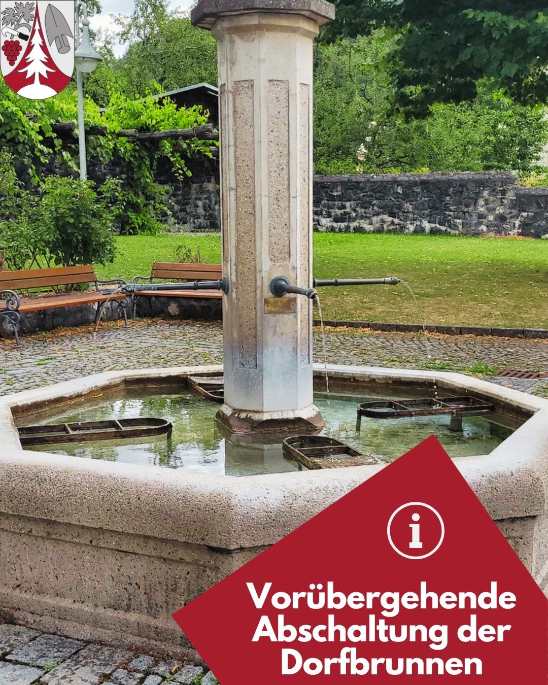 Ein steinerner Brunnen mit einem Wasserhahn in einem Park, umgeben von Bänken und Grün. Der Brunnen hat ein rotes Informationsschild.